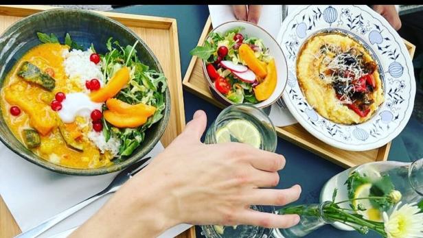 Verschiedene bunte Gerichte mit Salat, Suppe, Curry und Getränken werden auf Tabletts serviert, zwei Personen essen gemeinsam.