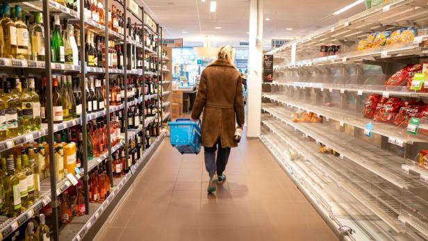 Person mit Einkaufskorb geht durch einen Supermarktgang mit leeren und vollen Regalen.