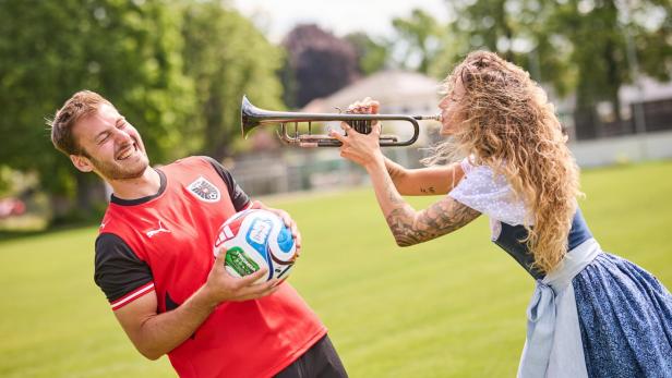 Ein lachender Mann im Fußballtrikot hält einen Ball, eine Frau in Tracht spielt ihm Trompete vor.
