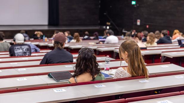Mehrere Studierende sitzen in einem Hörsaal mit nummerierten Plätzen und schauen zur Bühne nach vorne.