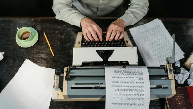 Person tippt an einer Schreibmaschine, umgeben von Papier, Bleistift und Kaffeetasse.