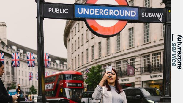Frau macht ein Selfie vor einem London Underground Schild, im Hintergrund rote Doppeldeckerbusse.