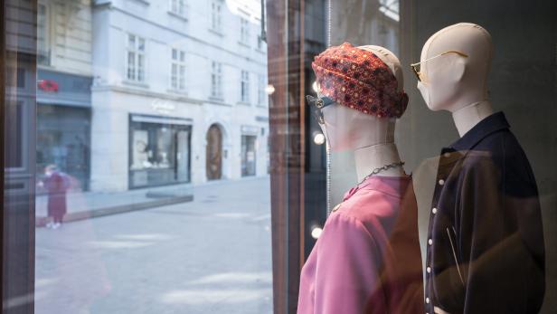 Zwei Schaufensterpuppen mit Sonnenbrillen und modischer Kleidung blicken durch ein Fenster auf eine ruhige Straße.