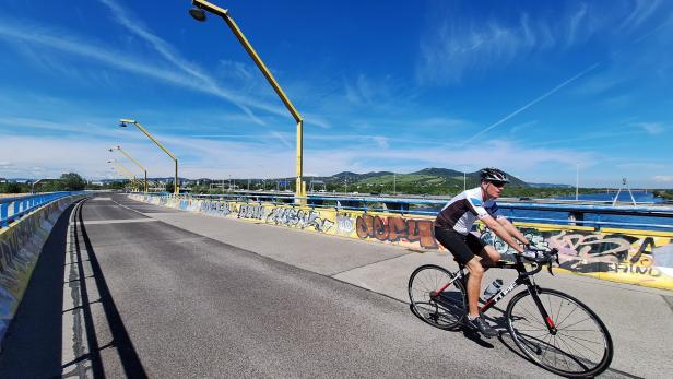 Ein Radfahrer fährt bei sonnigem Wetter auf einer mit Graffiti verzierten Brücke über einen Fluss.