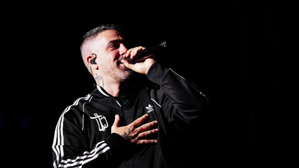 Bushido mit Tätowierungen singt mit Mikrofon in einer schwarzen Adidas-Jacke.