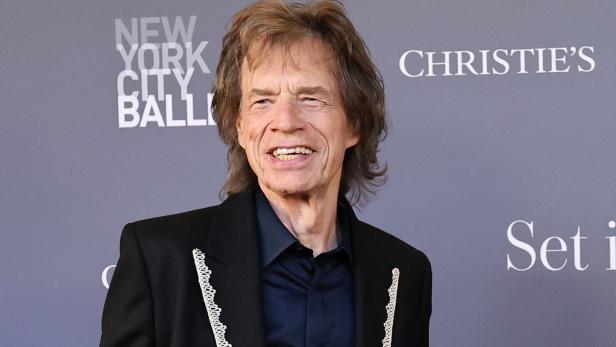 Mick Jagger lächelt vor einem grauen Hintergrund mit Logos.