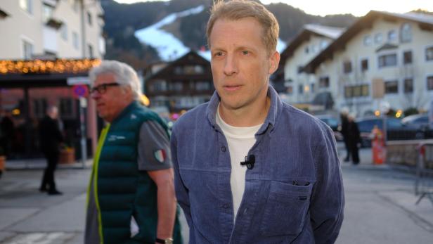 Oliver Pocher mit Mikrofon trägt ein blaues Cordhemd vor einem verschwommenen Hintergrund mit Häusern und Bergen.