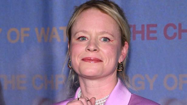 Thora Birch mit blonden Haaren trägt einen rosa Blazer und eine Perlenkette vor einem blauen Hintergrund.