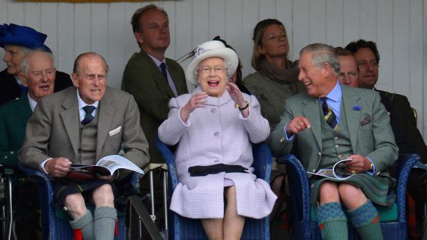 Queen Elizabeth II. und Charles lachen, während sie in traditioneller schottischer Kleidung auf Stühlen sitzen. Prinz Philip ist auch dabei.