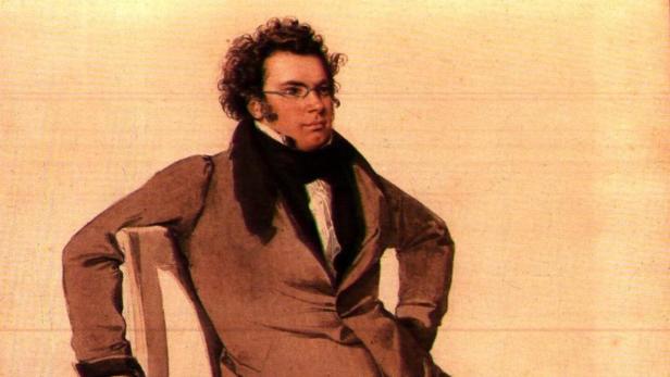 Franz Schubert sitzt lässig auf einem Stuhl, mit Brille und dunklem Mantel.