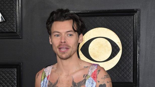 Harry Styles trägt einen bunten, rautenförmigen Overall und zeigt seine Tattoos.