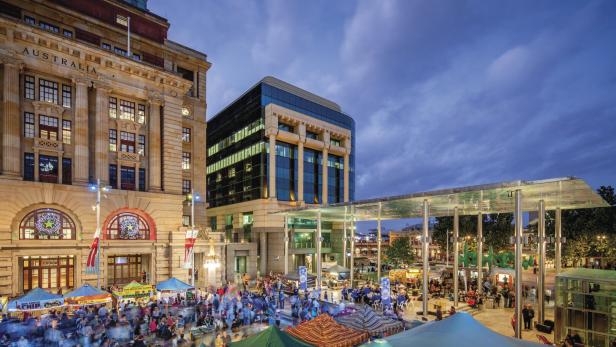 Ein belebter Marktplatz in Australien liegt zwischen historischen und modernen Gebäuden unter einem bewölkten Himmel.