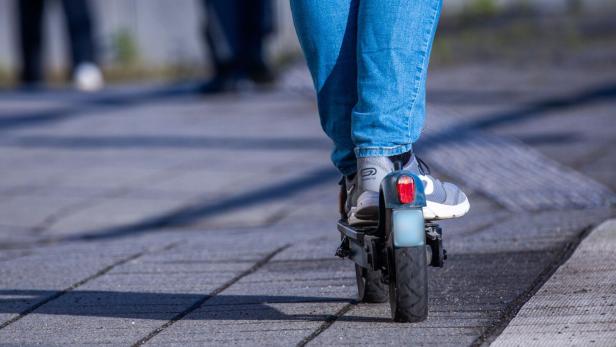 Person in blauer Jeans fährt auf einem E-Scooter auf einem gepflasterten Gehweg.