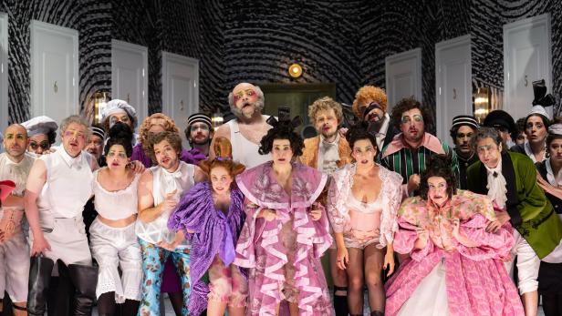 "Il viaggio a Reims" in Salzburg: Eine Gruppe von Darstellern in extravaganten Kostümen steht vor einer schwarz-weiß gemusterten Wand mit Türen.