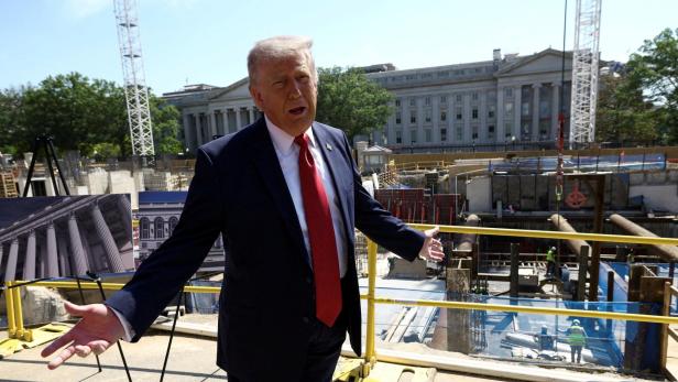 Donald Trump gestikuliert vor einer Baustelle mit einem großen Gebäude im Hintergrund.