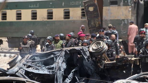 Nach einem Anschlag stehen Sicherheitskräfte vor einem zerstörten Auto, im Hintergrund ein Zug der Pakistan Railways.