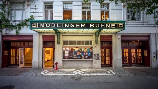 Eingang eines Theaters mit beleuchtetem Vordach, großen Fenstern und Plakaten an der Wand.