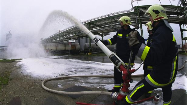 Feuerwehrleute spritzen mit einem Strahlrohr Löschschaum vor Industrieanlagen.