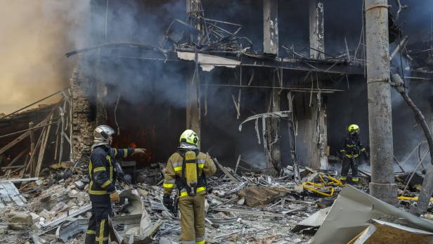 Kiew: Drei Feuerwehrleute stehen vor einem zerstörten, rauchenden Gebäude.