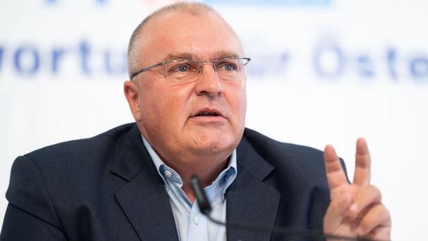 Ein Mann mit Brille und Anzug spricht und zeigt mit zwei Fingern eine Geste.