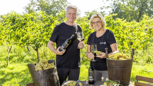 Zwei Personen stehen in einem Weinberg mit Weingläsern und Weinflaschen neben Körben voller Trauben.