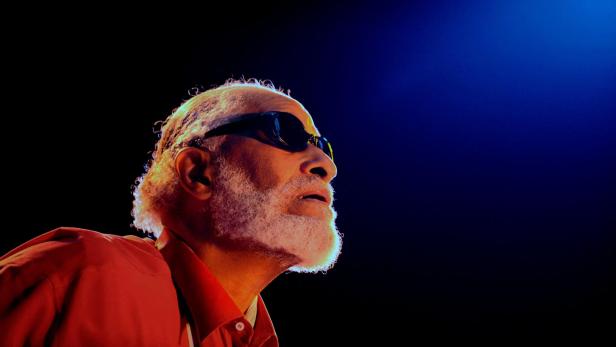 Ein Mann mit Sonnenbrille, rotem Hemd und weißem Bart blickt in ein blaues Licht.