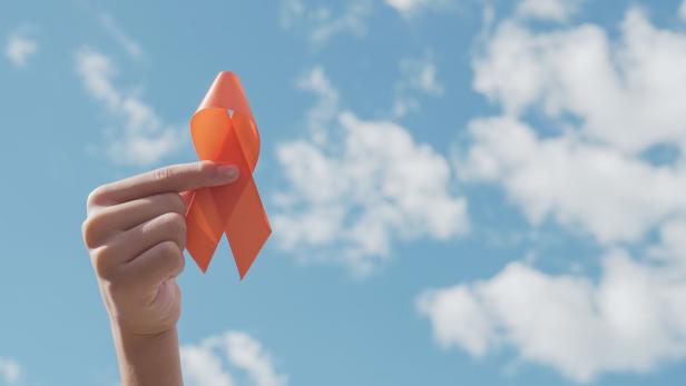Eine Hand hält eine orangefarbene Schleife vor blauem Himmel mit einigen weißen Wolken.