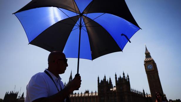 Ein Mann mit Sonnenbrille hält einen blau-schwarzen Regenschirm vor dem Big Ben und dem Palace of Westminster.