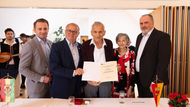 Fünf Personen stehen zusammen, eine Person in der Mitte hält eine Urkunde, im Hintergrund spielen Musiker, auf dem Tisch stehen Wasser und Blumen.