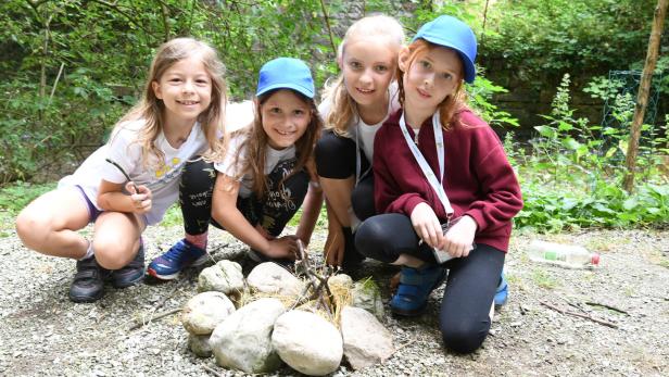 Vier Kinder hocken lachend um eine Feuerstelle aus Steinen im Grünen, zwei tragen blaue Kappen.