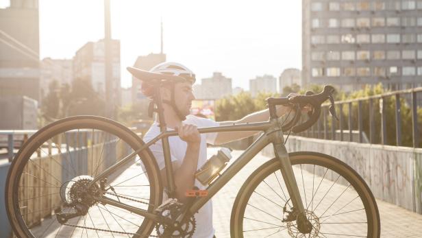 Ein Mann mit Fahrradhelm trägt ein Fahrrad auf der Schulter über eine Brücke in der Stadt bei Sonnenschein.