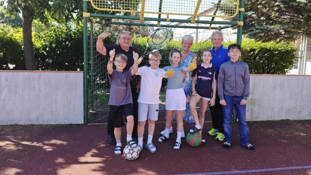 Sechs Kinder und drei Erwachsene stehen fröhlich auf einem Sportplatz vor einem Basketballkorb, einige halten Bälle in der Hand.