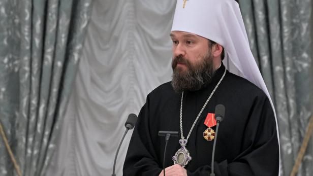 Ein bärtiger Mann in orthodoxer Kleidung und weißem Kopfschmuck mit Kreuz steht vor Mikrofonen.