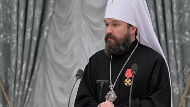 Ein bärtiger Mann in orthodoxer Kleidung und weißem Kopfschmuck mit Kreuz steht vor Mikrofonen.