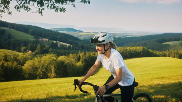 Eine Person mit Fahrradhelm fährt bei sonnigem Wetter durch eine grüne Hügellandschaft.