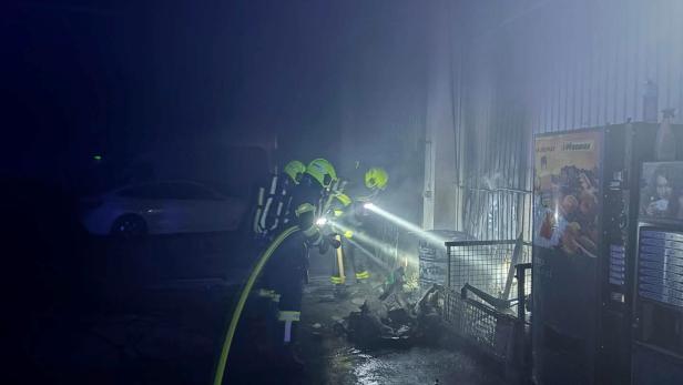 Zwei Feuerwehrleute mit Atemschutz löschen einen Brand in einer verrauchten Halle neben einem Getränkeautomaten.