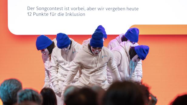 Eine Gruppe Tänzer mit weißen Masken und blauen Mützen performt vor Publikum.