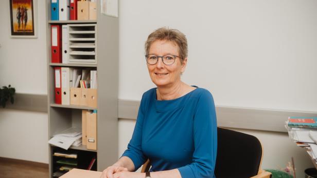 Eine Frau mit Brille und blauem Oberteil sitzt lächelnd an einem Schreibtisch in einem Büro mit Regal im Hintergrund.