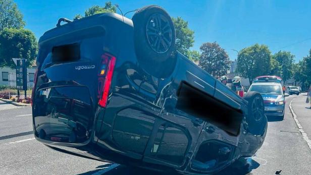 Ein Auto liegt auf dem Dach auf einer Straße, während Einsatzfahrzeuge und Personen im Hintergrund zu sehen sind.