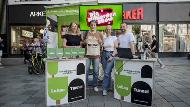 Vier Personen stehen an einem Stand mit der Aufschrift "Lobau Tunnel" und "Die Milliarden Show".