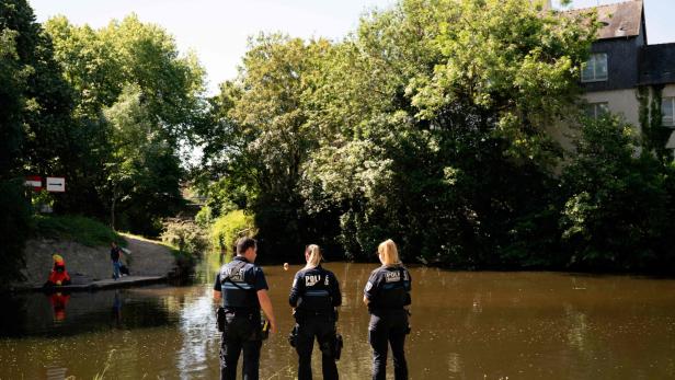 Drei Polizisten stehen am Ufer eines Flusses und blicken auf das Wasser, im Hintergrund sind Bäume und Häuser.