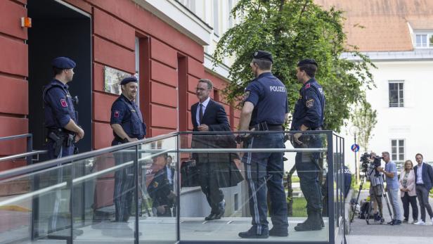 Ein Mann im Anzug geht an mehreren Polizisten vorbei, während im Hintergrund Kamerateams und Zuschauer stehen.