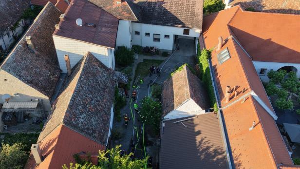 Mehrere Feuerwehrleute stehen in einem Innenhof zwischen Häusern mit roten Dächern und arbeiten mit Schläuchen und Leitern.