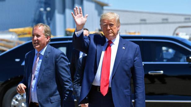 Donald Trump winkt in einem blauen Anzug mit roter Krawatte, begleitet von einem Mann im blauen Anzug.