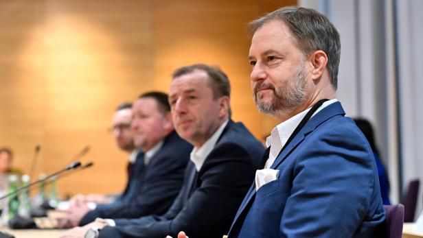 Vier Männer in Anzügen sitzen nebeneinander an einem Konferenztisch und hören aufmerksam zu.