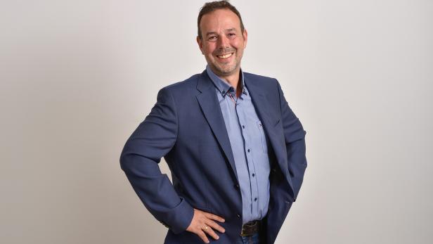 Ein Mann im blauen Anzug steht mit den Händen in die Hüften gestützt und lächelt freundlich vor hellem Hintergrund.
