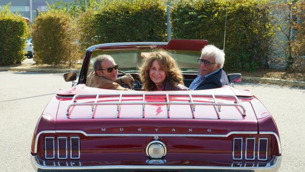 Antoine, Victoire und der Manager sitzen im offen Cabrio und schauen zurück.