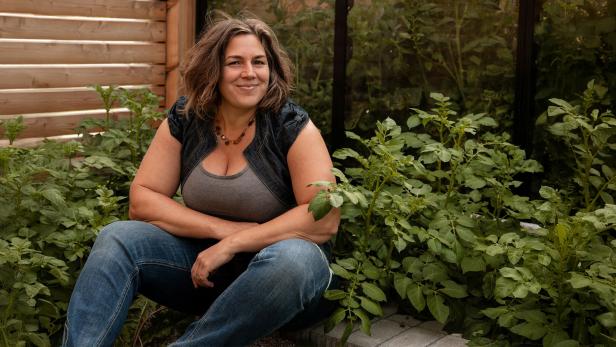 Eine Frau sitzt lächelnd in einem Gartenbeet, umgeben von üppigen grünen Pflanzen und trägt legere Kleidung.