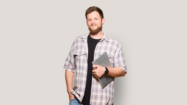 Ein junger Mann mit Bart und kariertem Hemd hält einen Laptop und lächelt vor hellem Hintergrund.