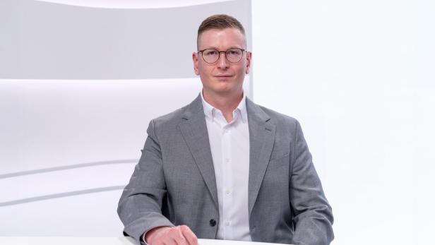 Mann mit Brille und kurzem Haar trägt grauen Anzug und weißes Hemd, sitzt an einem weißen Tisch vor hellem Hintergrund.
